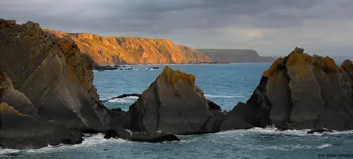 This landscape photograph captures the Cornish cliffs along the coast of Cornwall in the United Kingdom, with dramatic rock formations rising from the sea. The image was taken during the evening in autumn, as suggested by the warm, low-angle sunlight illuminating the rugged cliff faces and the rich golden hues spreading across the rural landscape. The natural coastline, shaped by years of erosion, forms distinctive triangular shapes in the foreground, and the calm sea contrasts with the imposing cliffs behind. This scene exemplifies the unspoiled nature of the area, highlighting the wild beauty of rural Cornwall by the sea.